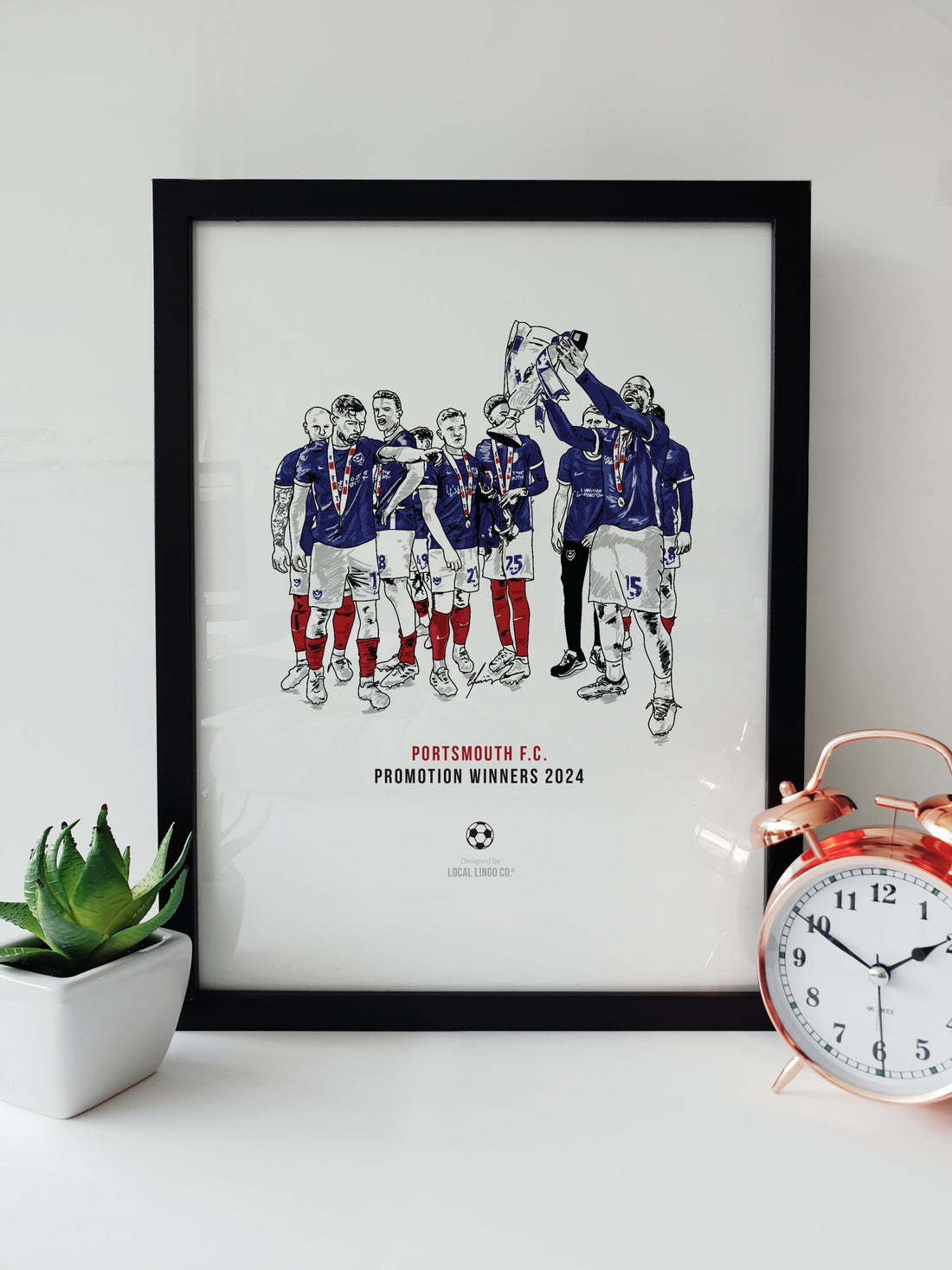 Framed Portsmouth FC Promotion Winners Print featuring a team celebration illustration, displayed on a white desk with a plant and a copper alarm clock. designed by local lingo