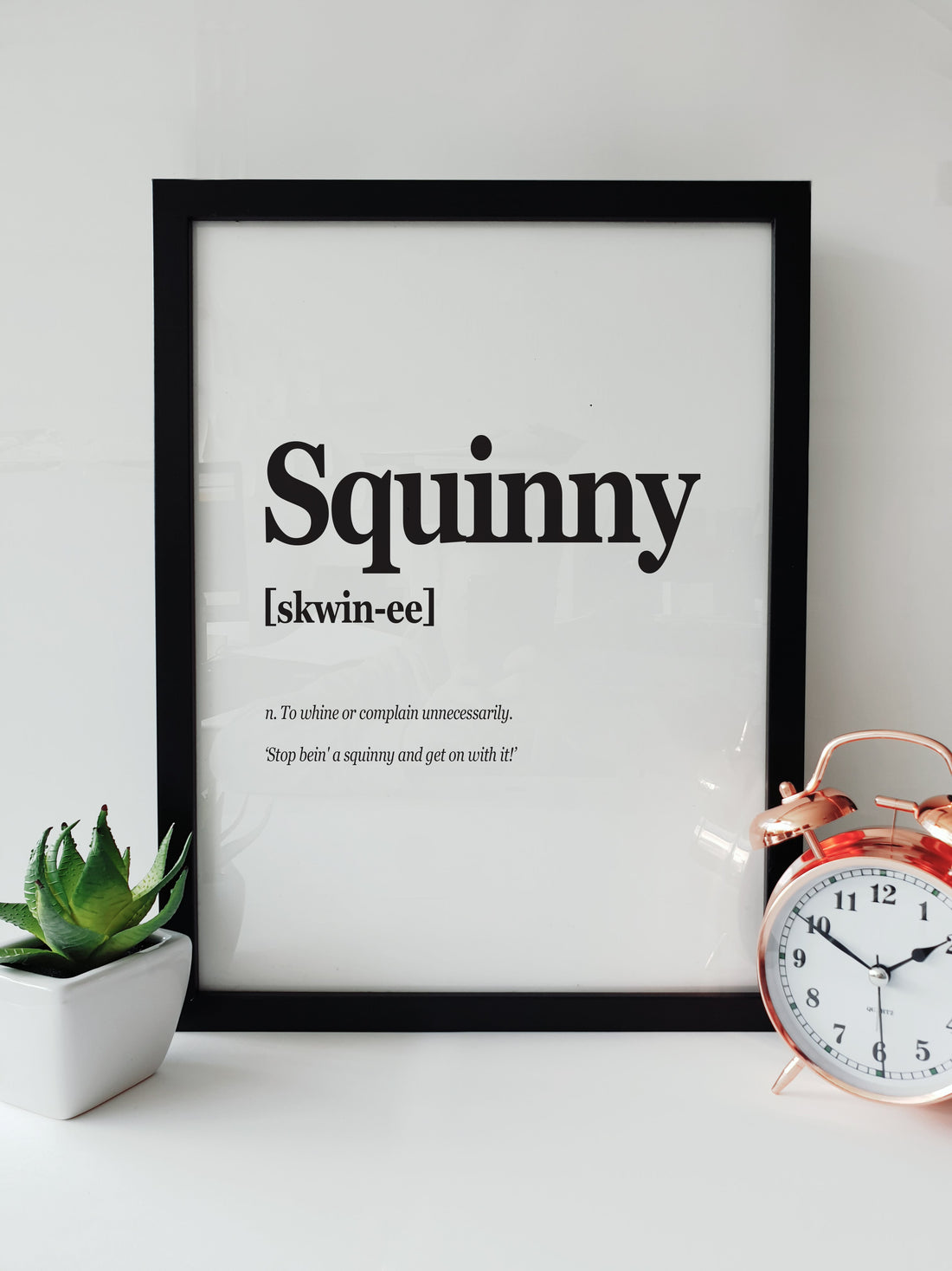 Framed Portsmouth Dialect Print featuring the word "Squinny" with pronunciation and definition, displayed on a white desk with a plant and a copper alarm clock. pompey translation designed by local lingo